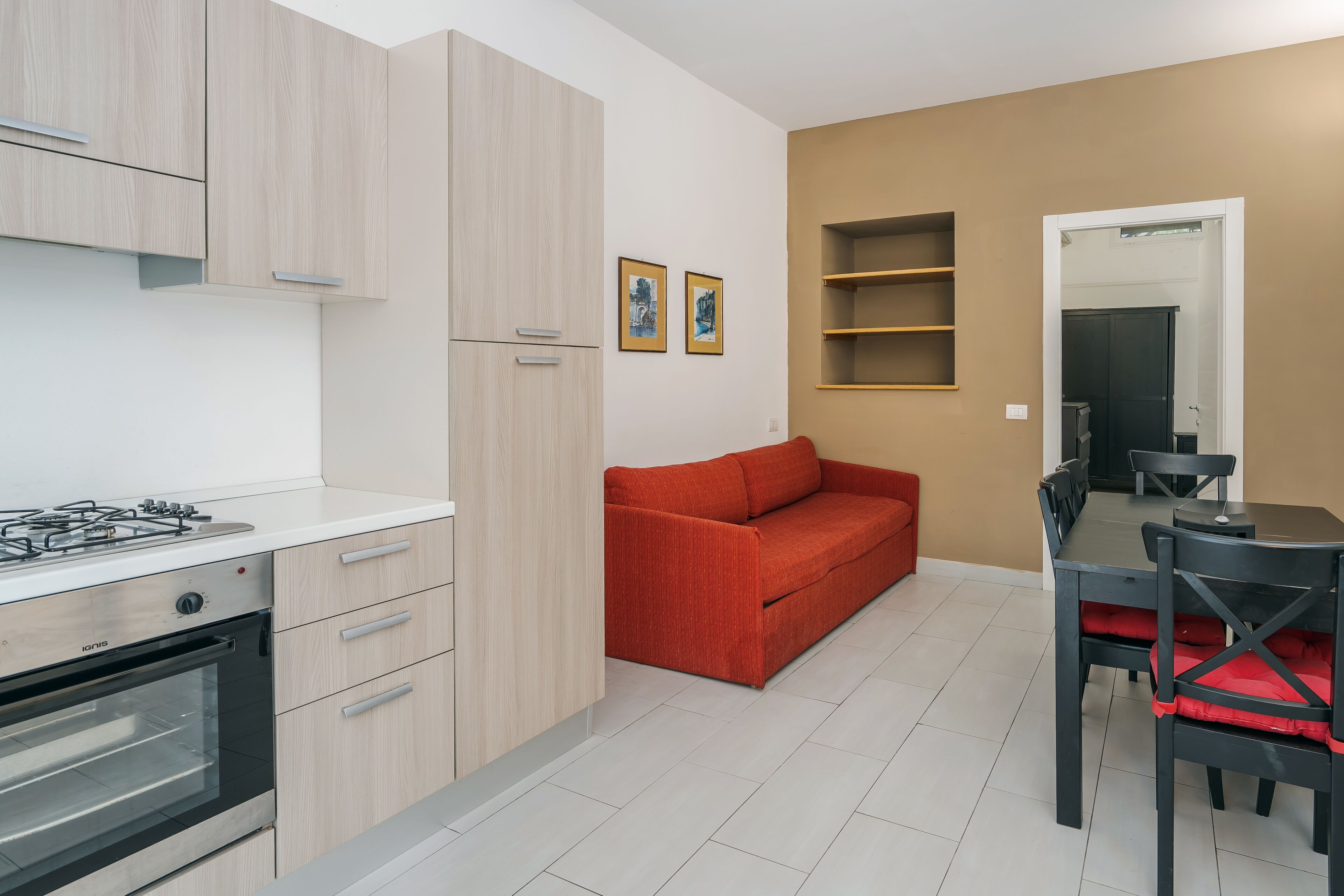 A modern kitchen with a gas stove, oven, and light wood cabinets. There is a red sofa and a dining table with chairs in the living area.