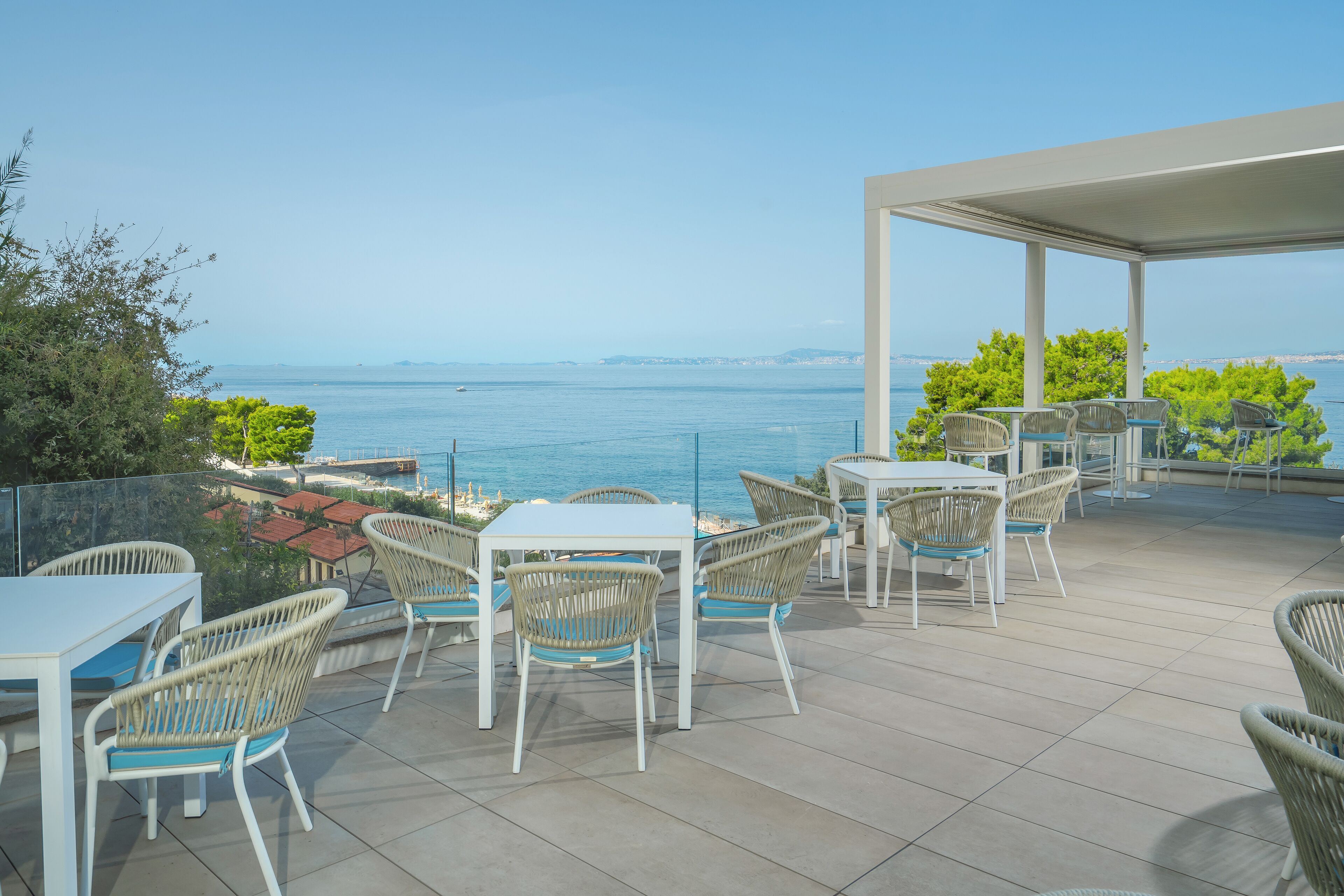 An outdoor patio with white tables and wicker chairs, overlooking a body of water and distant land.