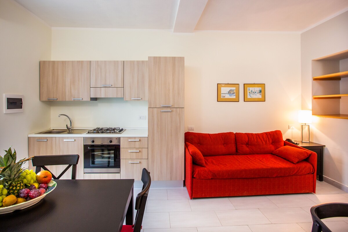 A modern kitchen with light wood cabinets, a dining table with chairs, and a red sofa.