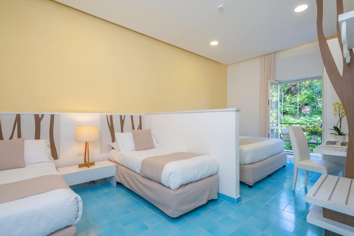 A hotel room with two beds, a desk, and a chair. There is a window with a view of greenery.