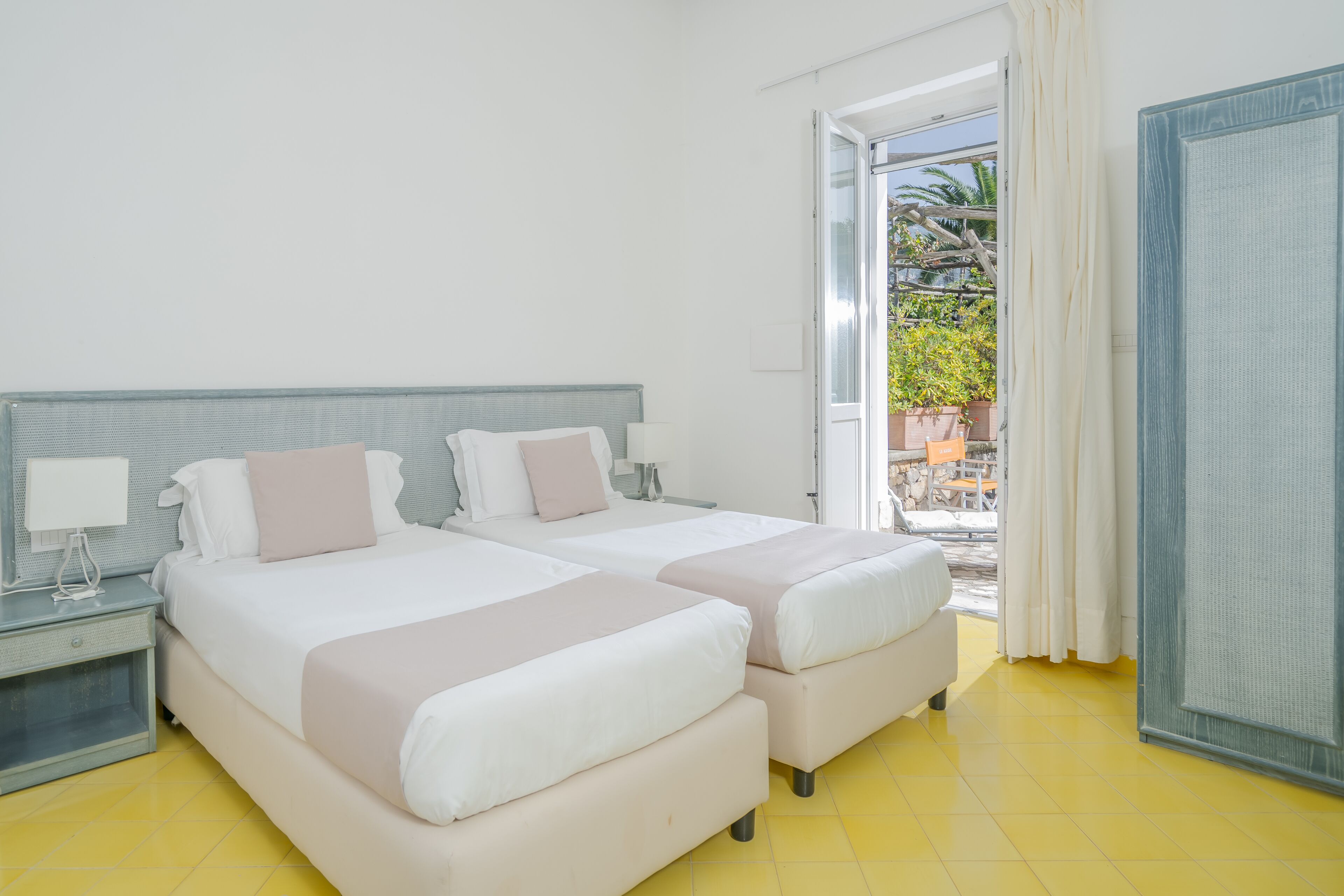 A hotel room with two beds, a nightstand, a window with curtains, and a view of the outside.