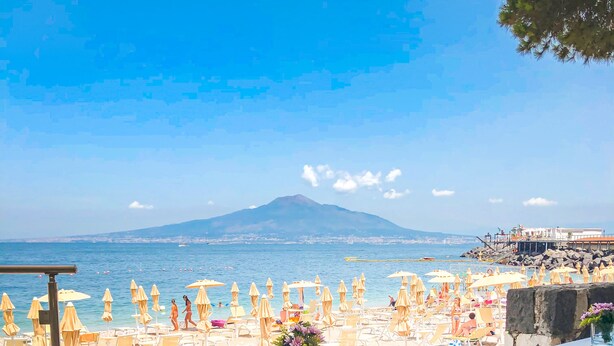 An outdoor seating area with a table and chairs, overlooking a beach with umbrellas and people. - Hotel & Resort Le Axidie (Vico Equense)