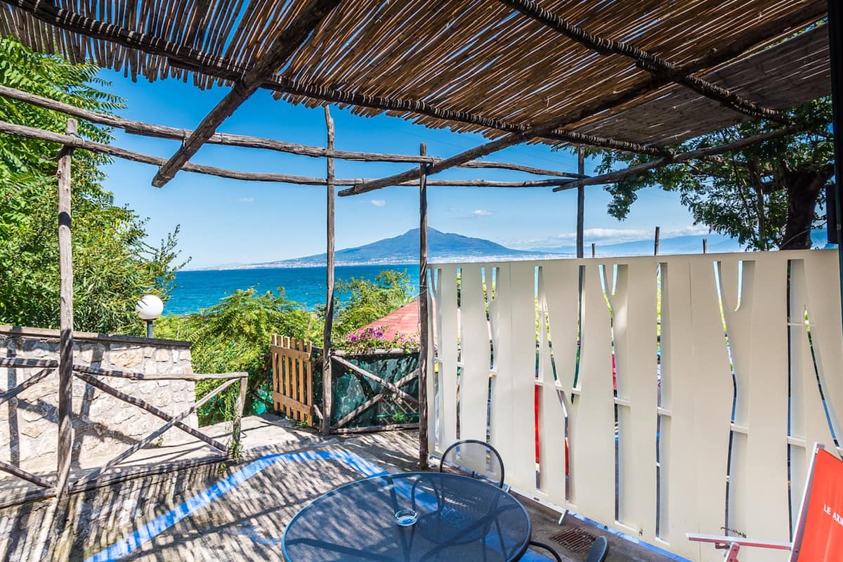 A covered outdoor seating area with a table and chairs, overlooking a scenic view of the sea and a mountain.