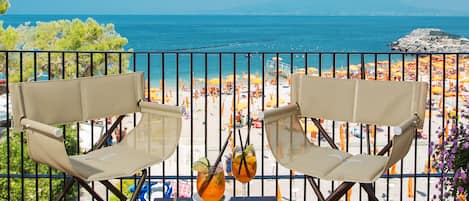 A balcony with two chairs and a table, overlooking a beach with umbrellas and people.