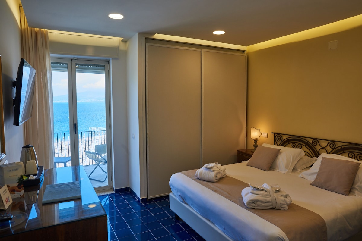 A hotel room with a bed, a desk with a computer, a TV, and a view of the sea through a sliding door.