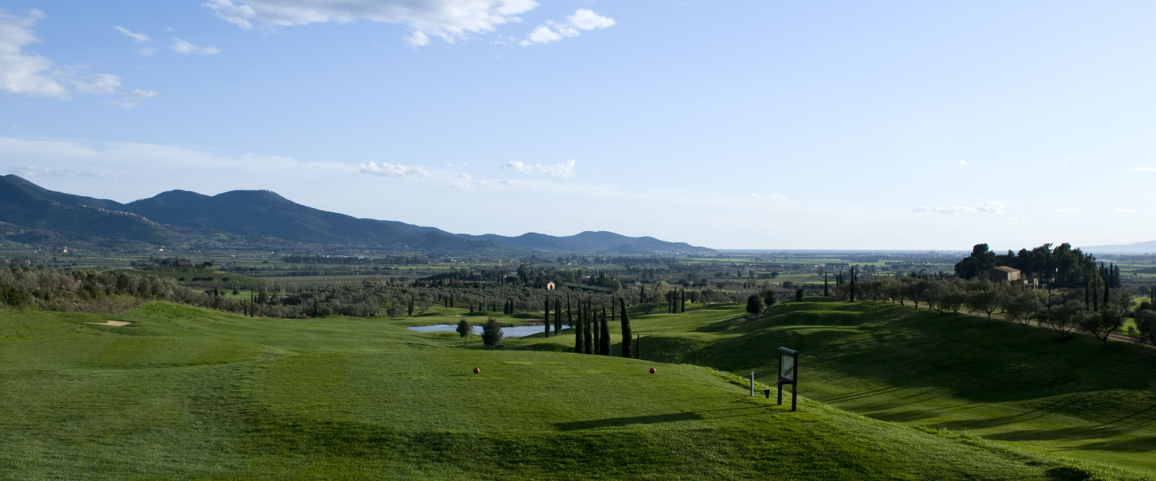A golf course with rolling hills, a pond, and distant mountains.