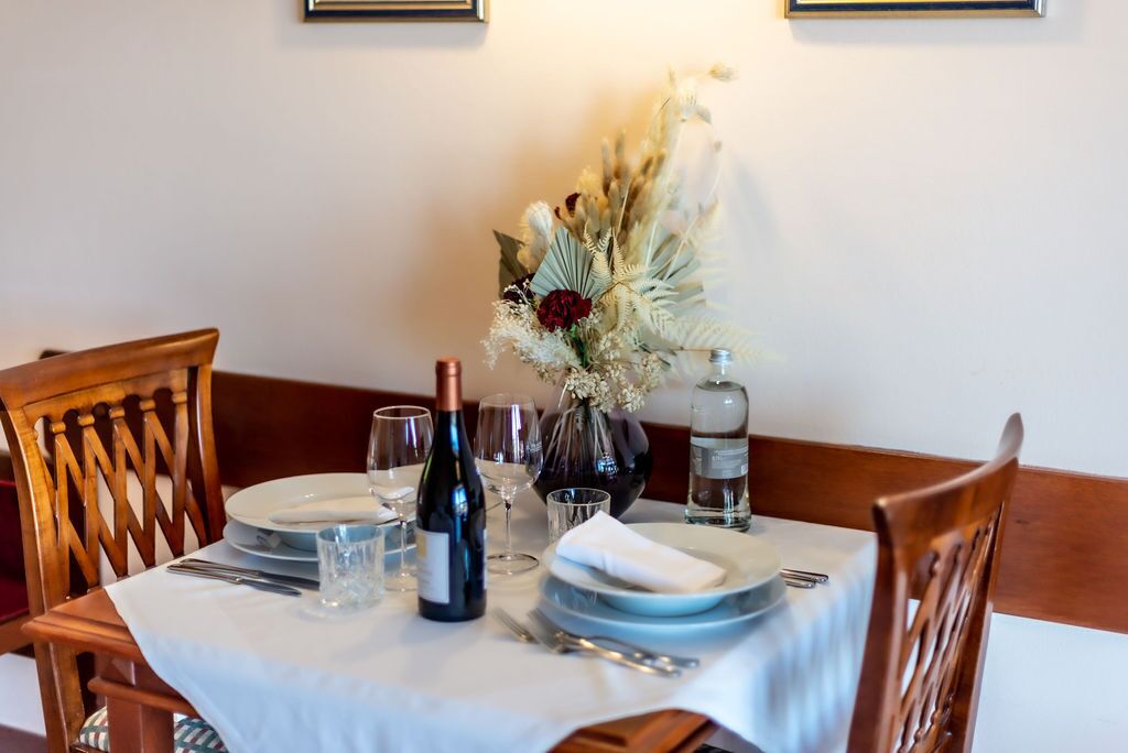 Una mesa de comedor dispuesta con platos, cubiertos, copas de vino y un florero con flores secas.