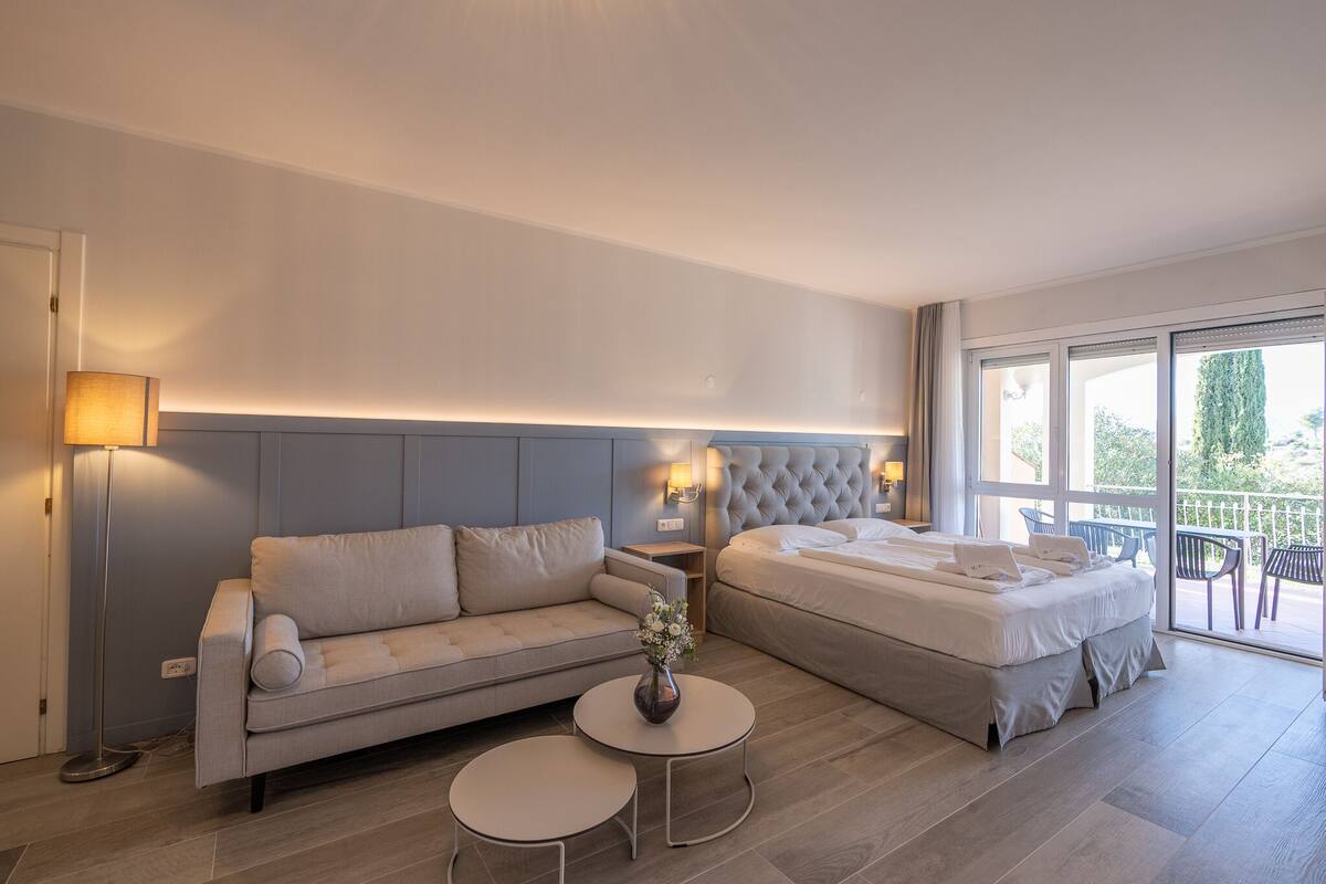 Ein modernes Hotelzimmer mit einer Couch, einem Bett, einem kleinen Tisch mit einer Vase und einem Balkon mit Stühlen.