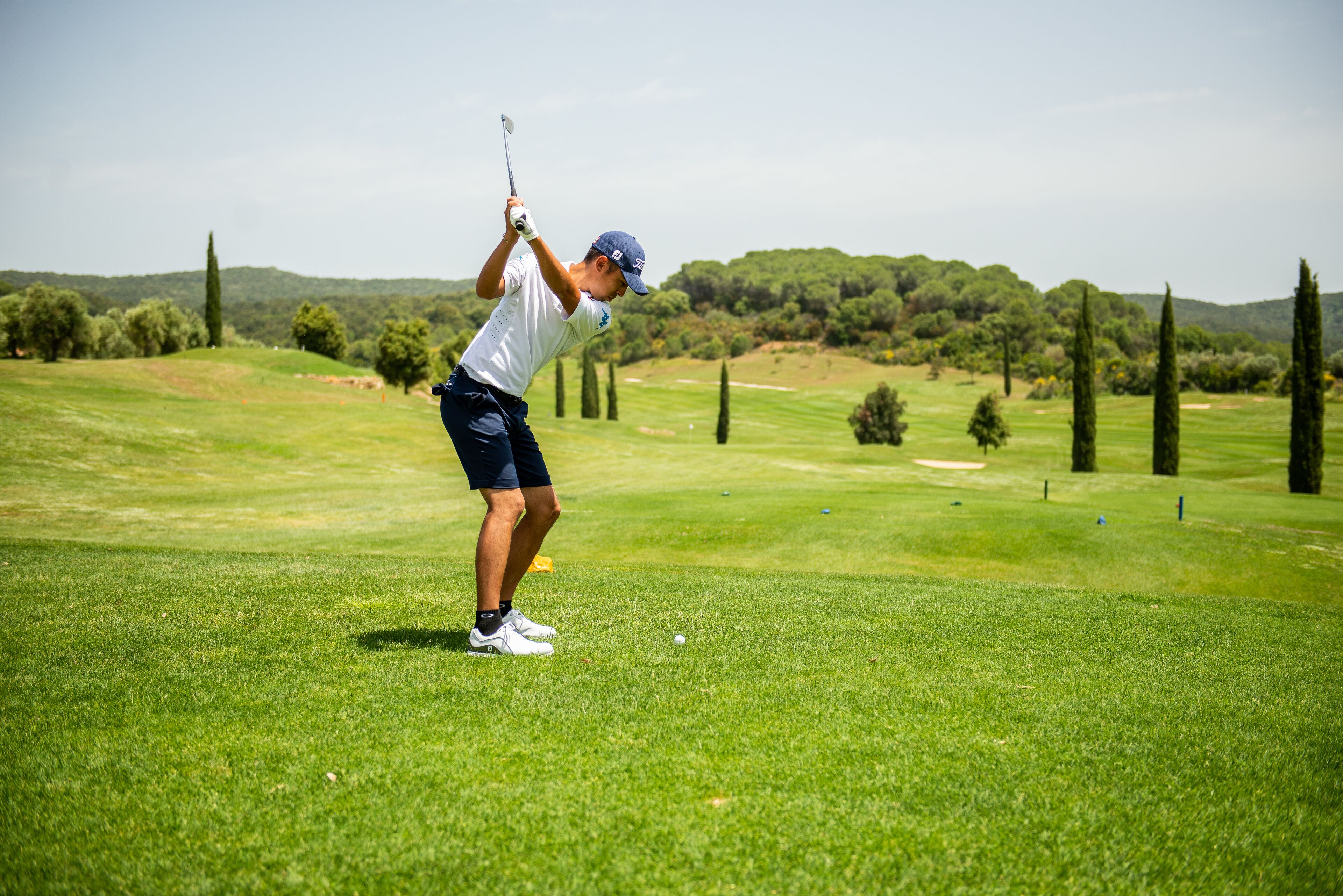 A golf course with a player in mid-swing, a well-maintained green, and a backdrop of trees and hills.