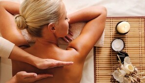Une personne reçoit un massage du dos dans un spa, avec une serviette, des bougies et un petit pot de crème à proximité.
