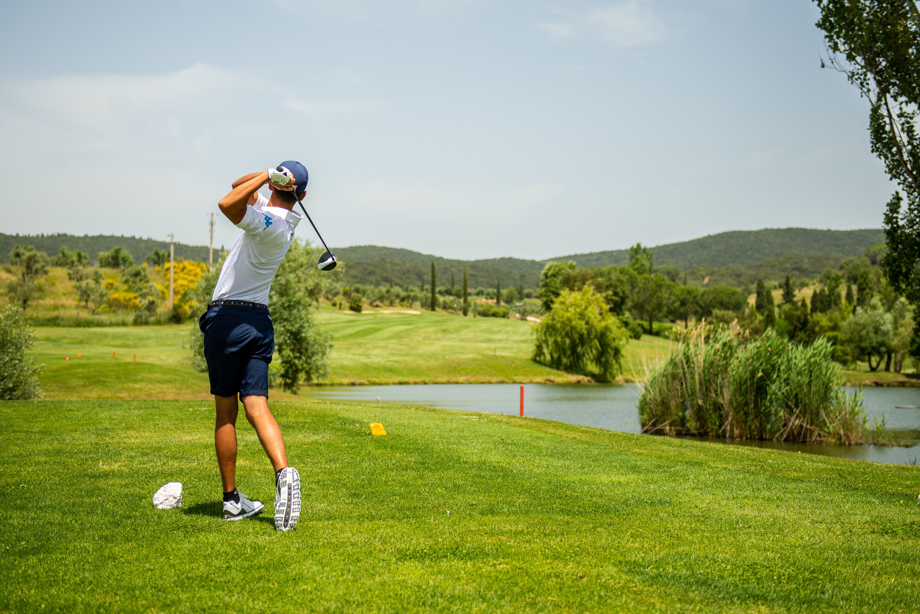 Een golfbaan met een speler in een wit shirt en blauwe korte broek, die zich klaarmaakt om een bal te slaan.