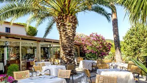 Un área de comedor al aire libre con mesas dispuestas para dos, rodeada de palmeras y plantas en flor.