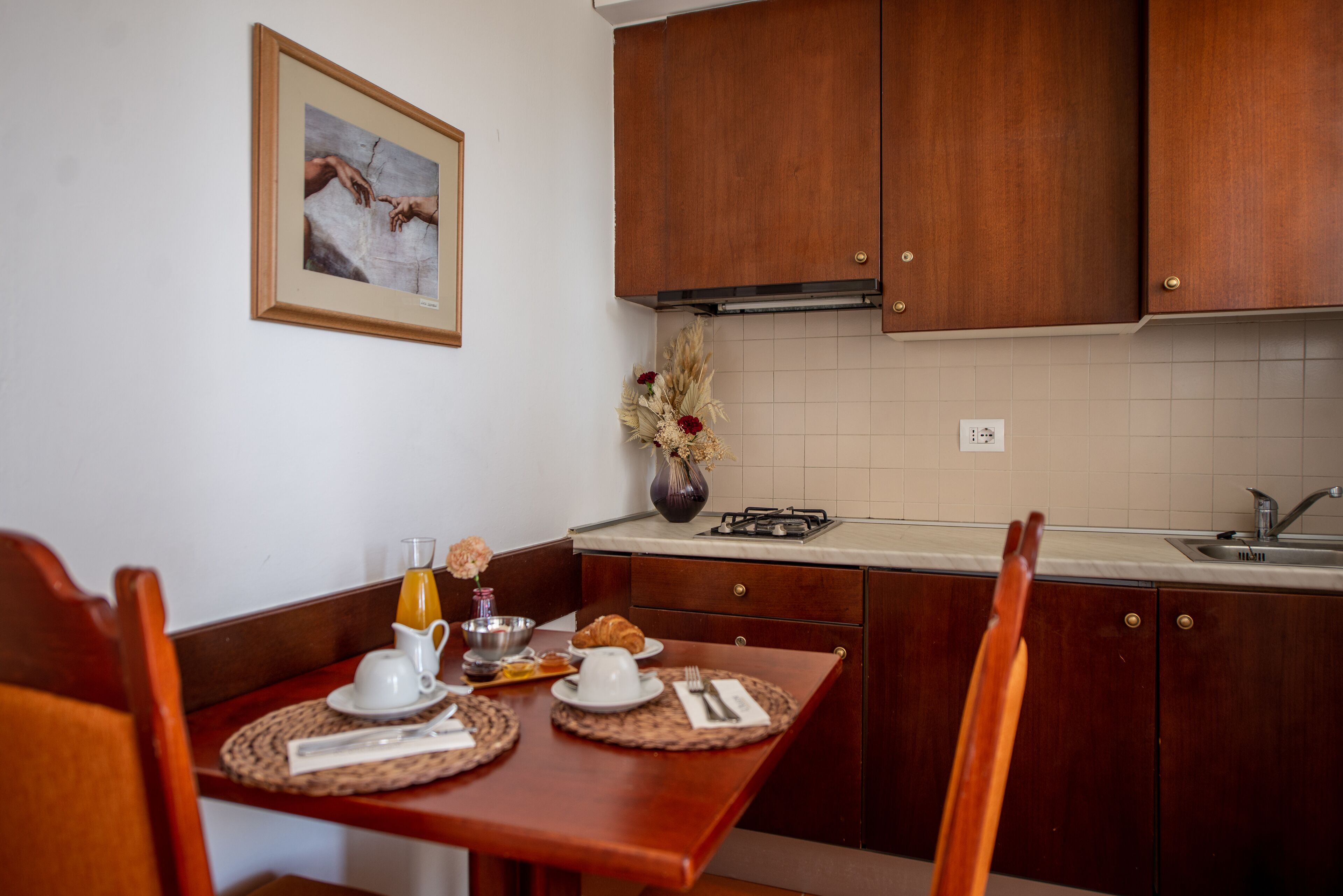 Une cuisine avec des meubles de rangement en bois, une table à manger avec des sets de table, un vase contenant des fleurs séchées et un tableau encadré accroché au mur.