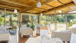 Un área de comedor al aire libre cubierta con mesas dispuestas para una comida, rodeada de paredes de vidrio y vigas de madera.