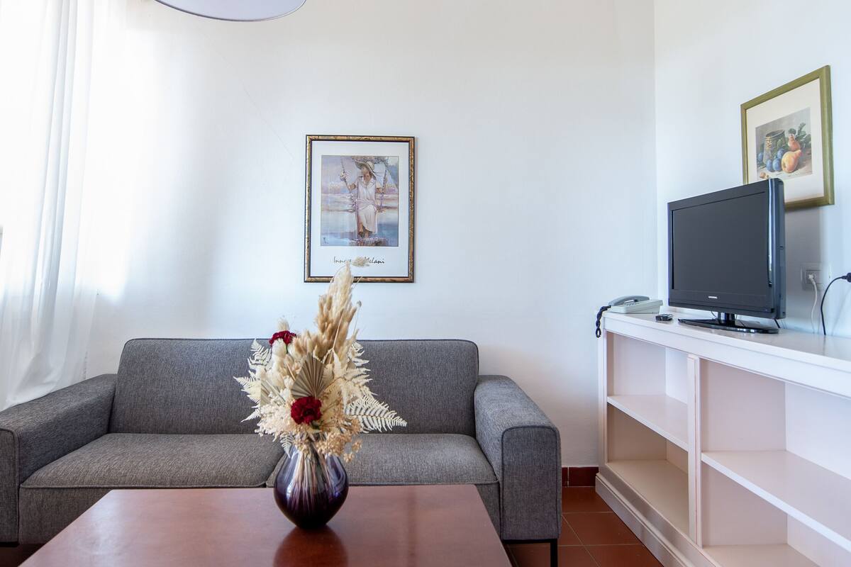 Una sala de estar moderna con un sofá gris, una mesa de centro de madera, un televisor sobre un mueble bajo y una lámpara en el techo.