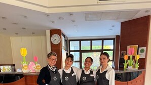 Un grupo de personas en el vestíbulo de un hotel, con una mesa de recepción, un reloj y un ventanal con vistas a zonas verdes.