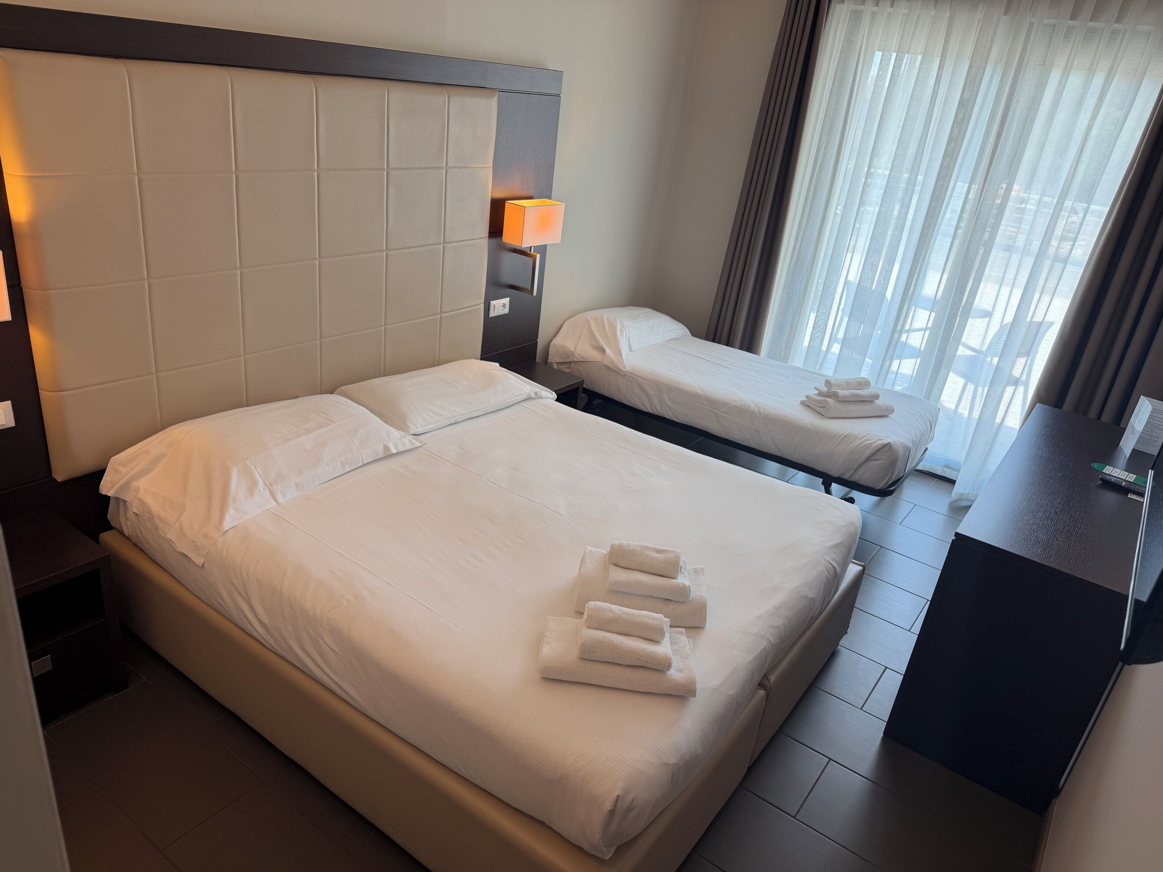 Een hotelkamer met twee bedden, een nachtkastje, een lamp en uitzicht op de stad door het raam.