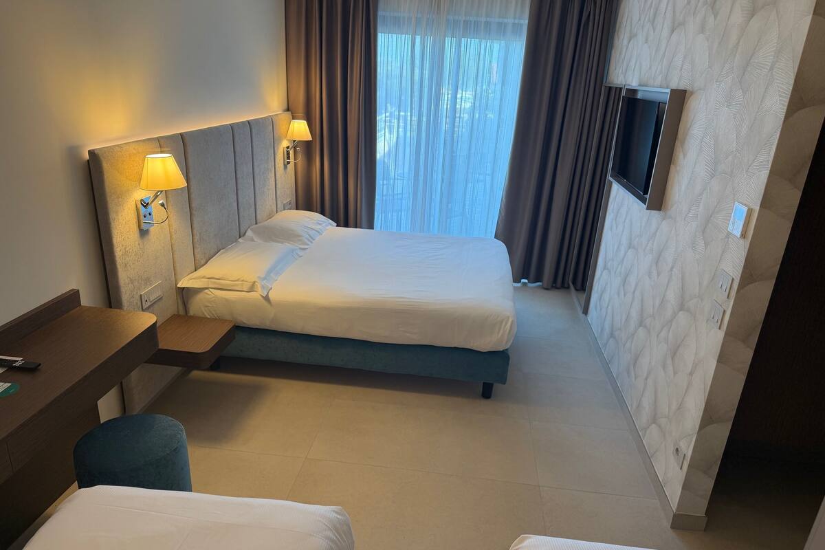 Quarto de hotel com cama, mesas de cabeceira, escrivaninha, cadeira, televisão e cortinas.