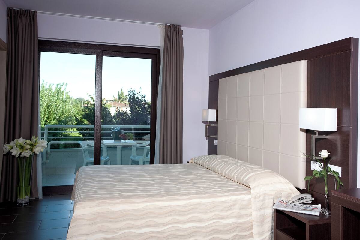 Quarto de hotel com uma cama, mesa de cabeceira com abajur, vaso com flores e vista para uma varanda com mobiliário.