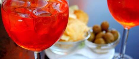 Dos vasos de bebida roja con cubitos de hielo, un cuenco de aceitunas y un plato de aperitivos.