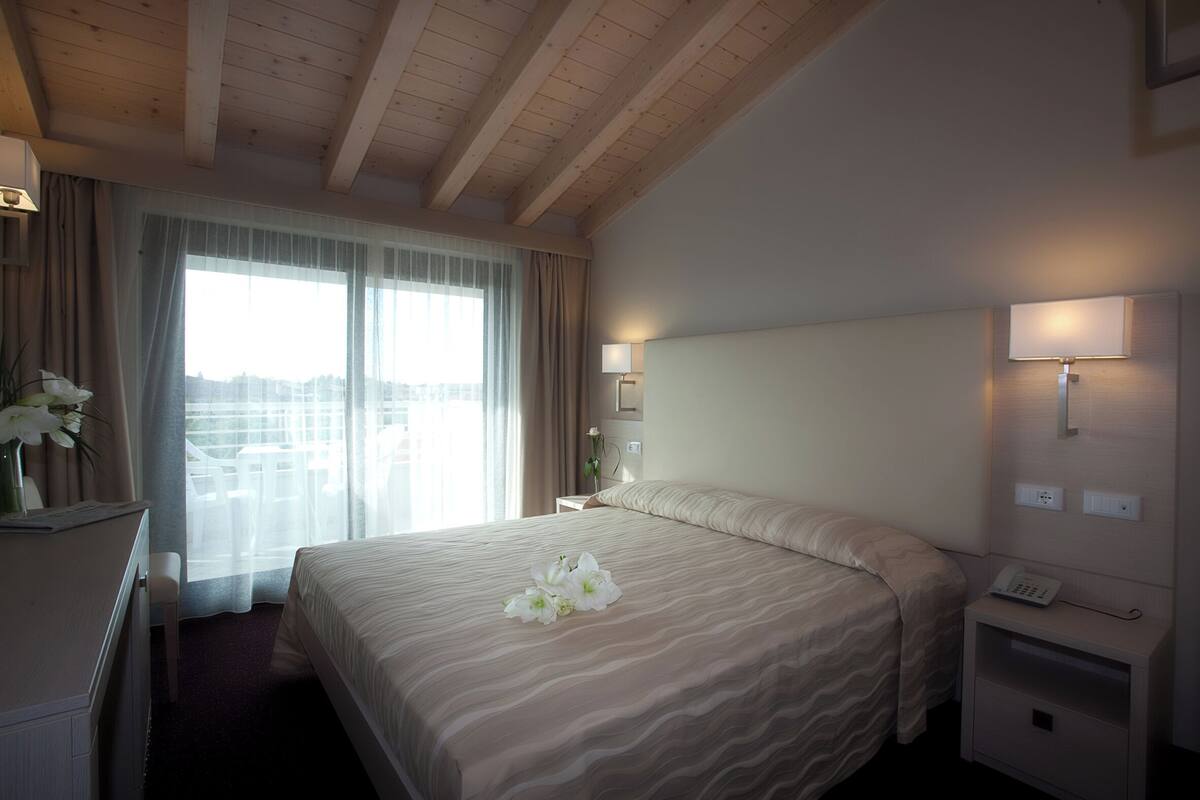 Quarto de hotel com uma cama grande, uma mesa de cabeceira com abajur, um criado-mudo com telefone e uma janela com cortinas.