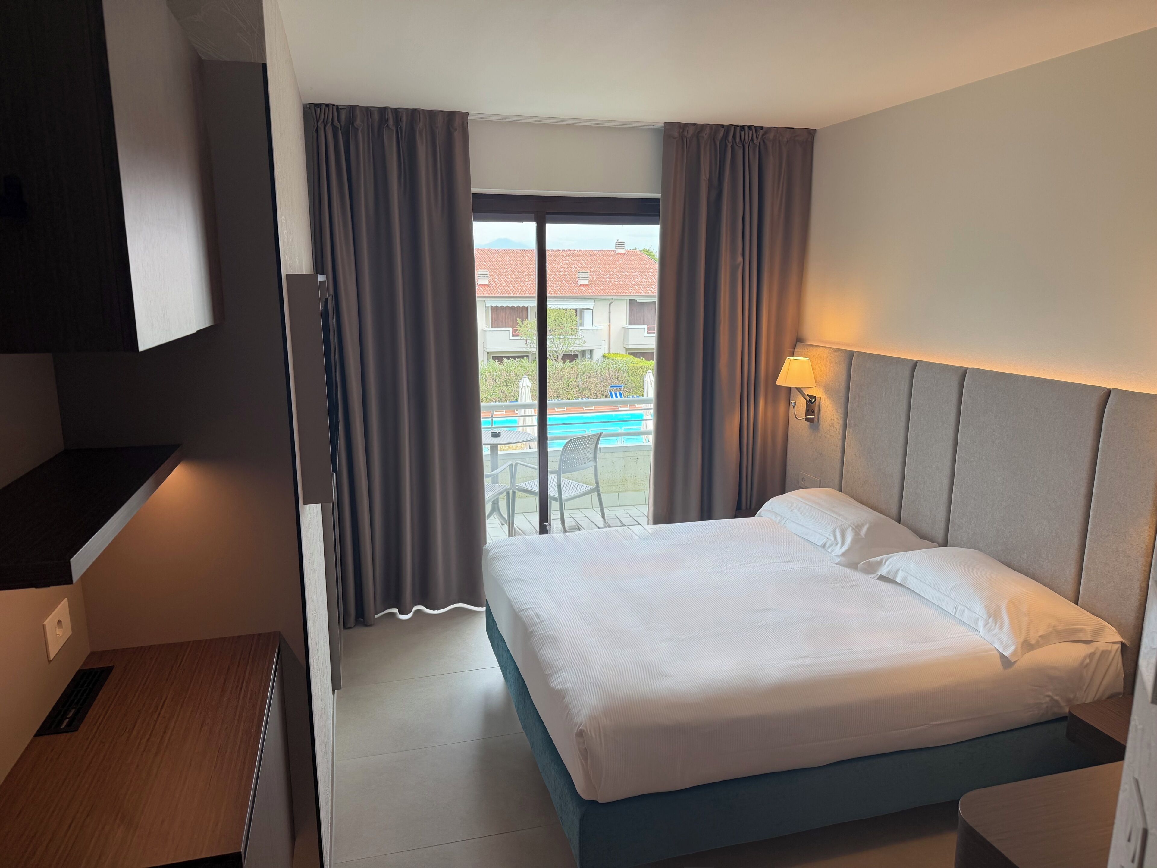 A hotel room with a bed, a desk, a chair, a lamp, and a view of a swimming pool.