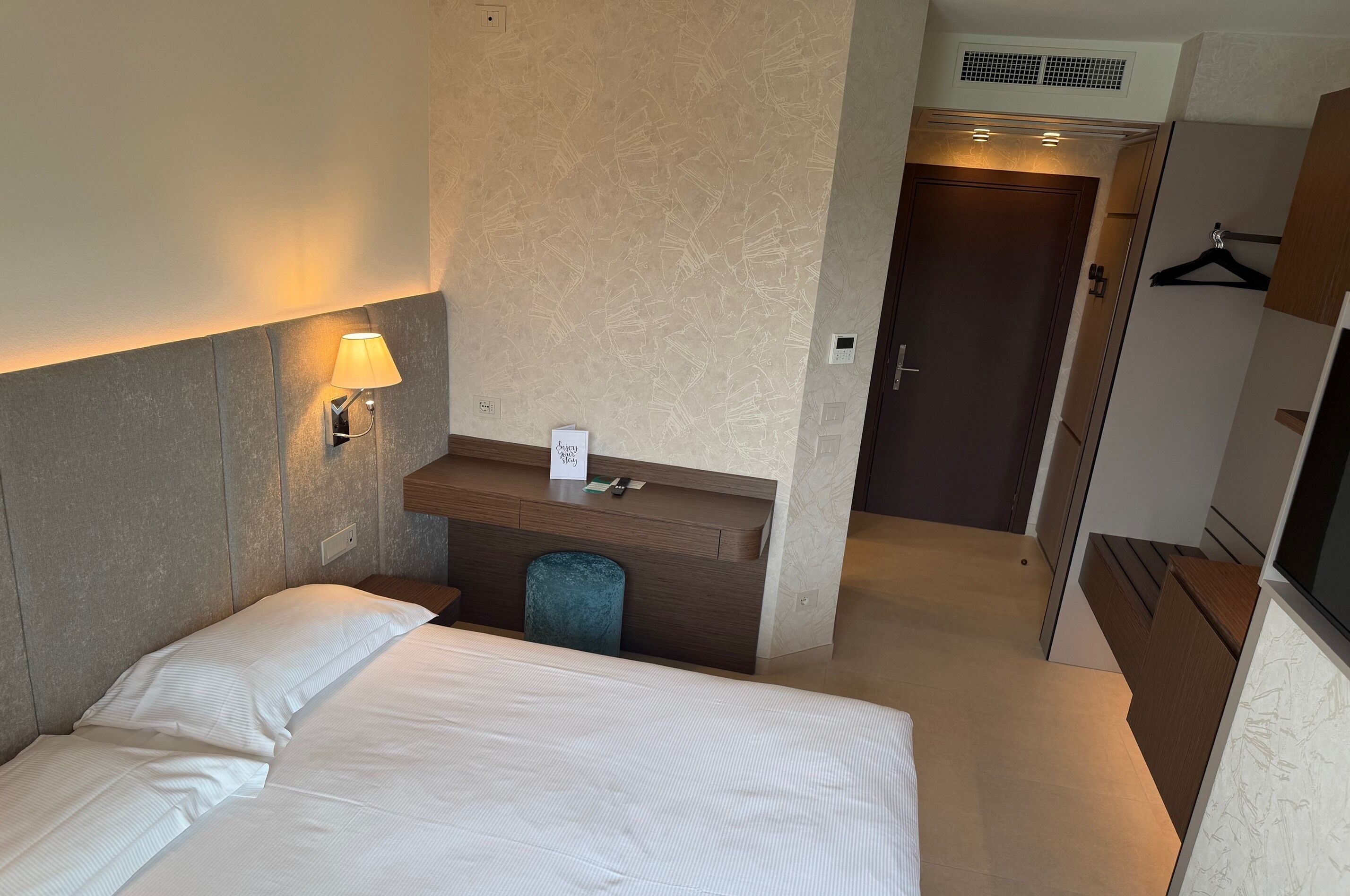 A hotel room with a bed, bedside lamp, a small desk with a chair, a coat rack, and a door leading to another room.