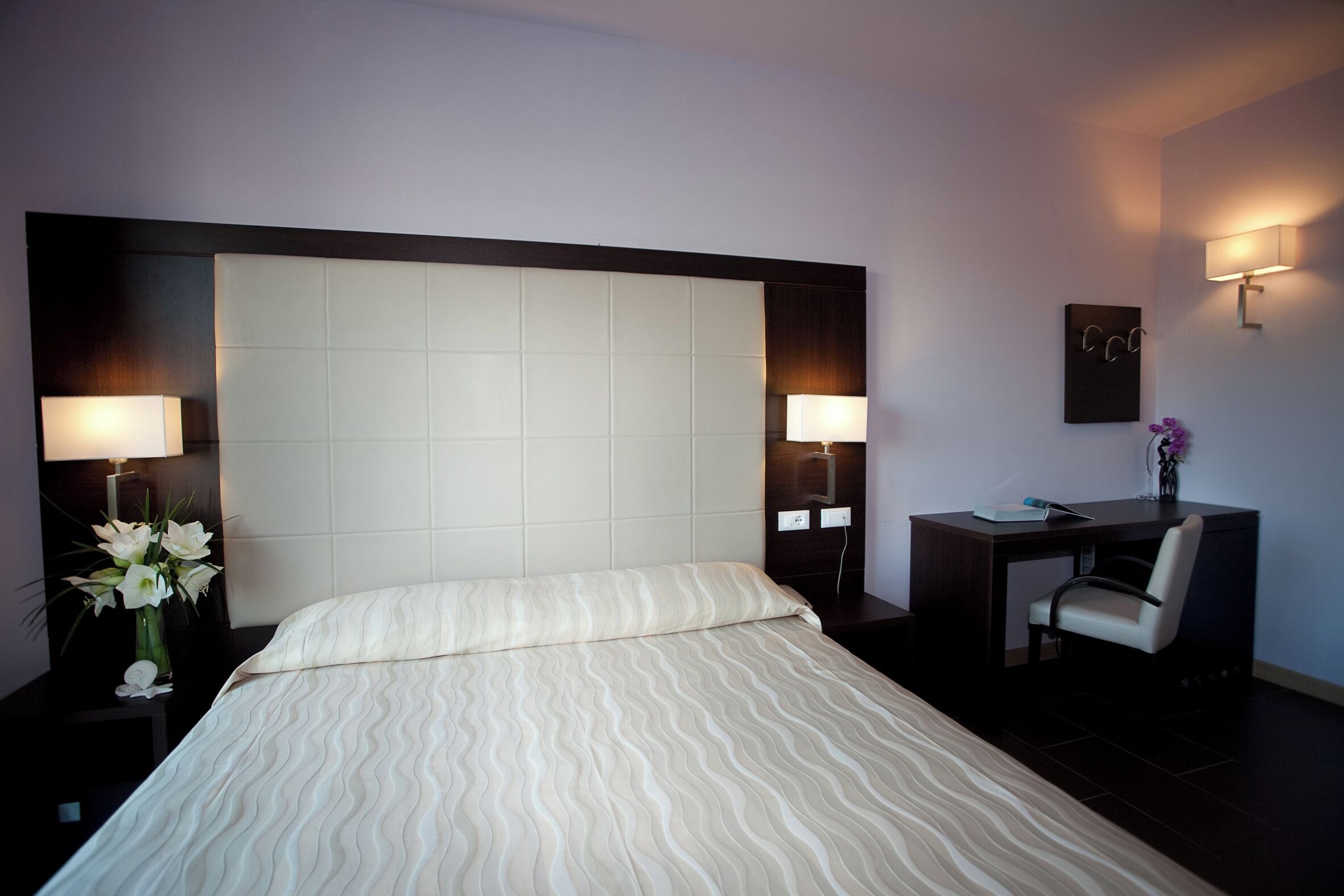 A hotel room with a large bed, two bedside lamps, a desk with a chair, and a small vase with flowers.