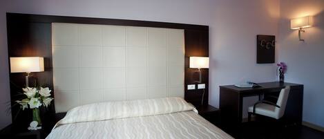 Habitación de hotel con una cama grande, dos lámparas de noche, un escritorio con silla y un pequeño jarrón con flores.