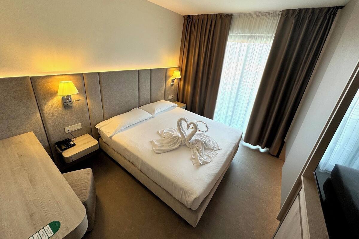 Quarto de hotel com cama, abajures, mesa de cabeceira, telefone e janela com cortinas.