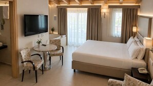 Una habitación de hotel con una cama, un televisor, una mesa redonda con sillas, una mesita con una planta y una ventana con cortinas.