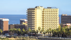 Un hotel de varios pisos con balcones, situado cerca de una playa con palmeras.