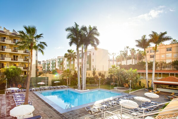 Una piscina rodeada de sillas de descanso y sombrillas, con palmeras y un edificio al fondo.