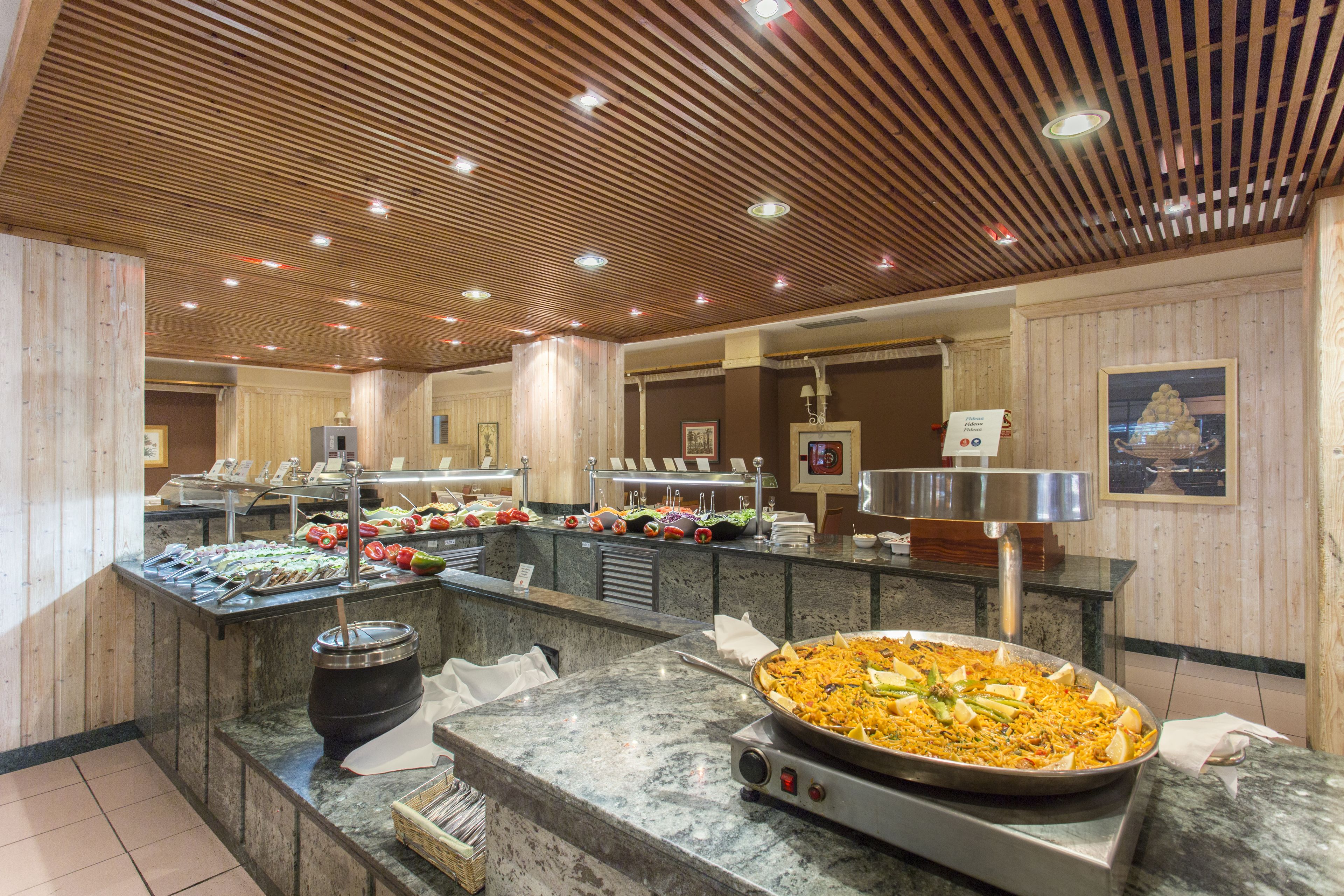A buffet area with a large pan of food, multiple serving stations, and a wooden ceiling.