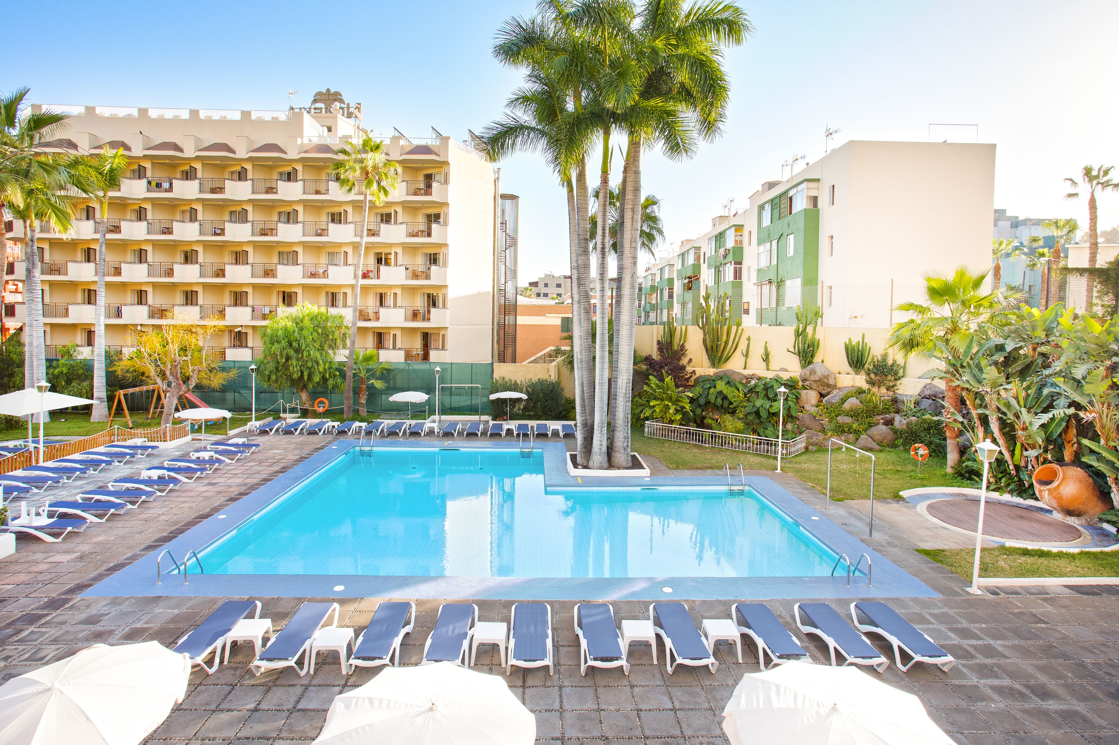 A swimming pool with sun loungers and umbrellas, surrounded by palm trees and a multi-story building.