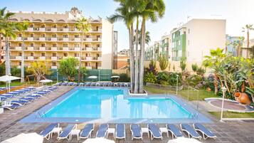 A swimming pool with sun loungers and umbrellas, surrounded by palm trees and a multi-story building.