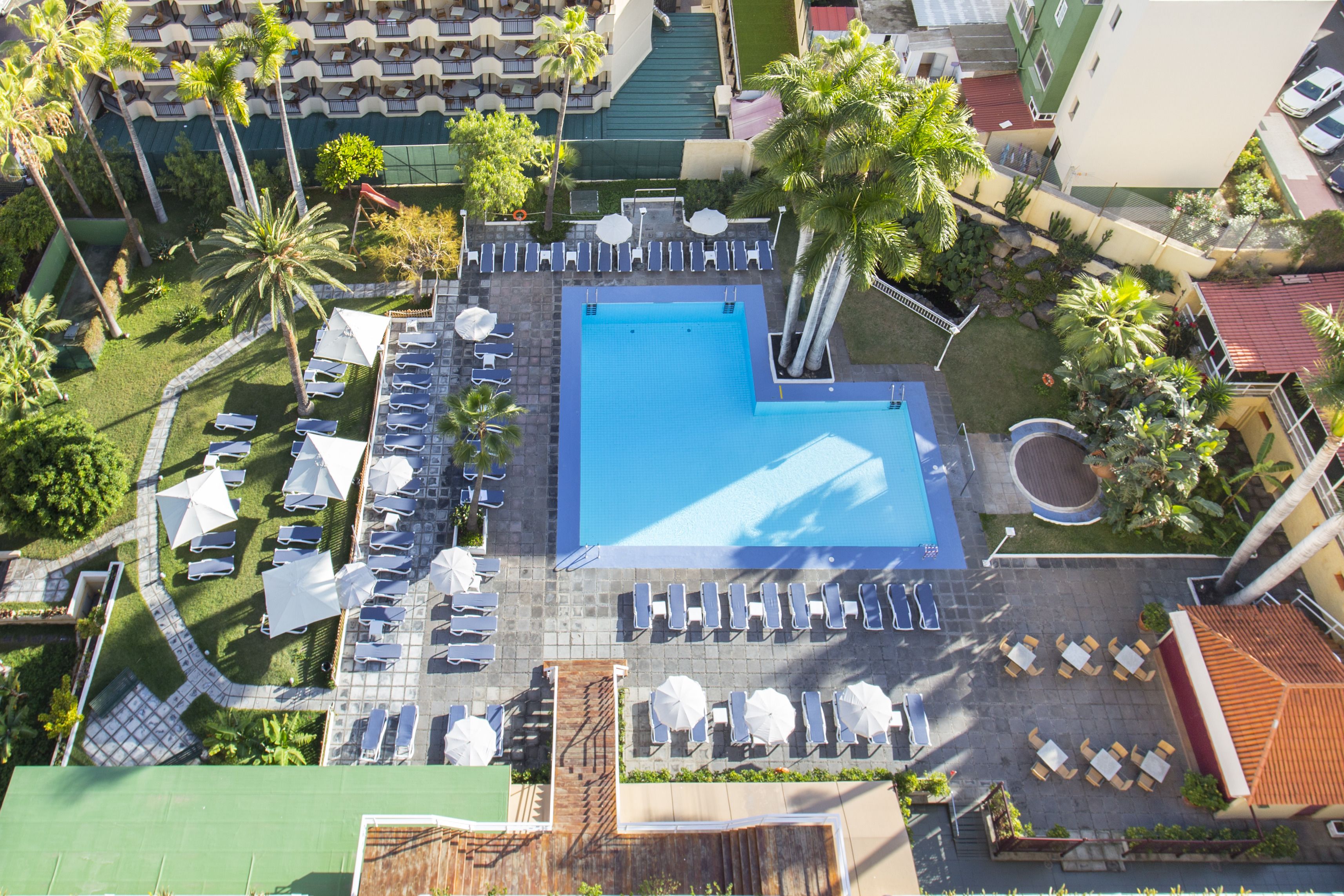 An aerial view of a hotel with a central swimming pool surrounded by sun loungers and umbrellas, a small garden area, and a parking lot.