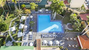An aerial view of a hotel with a central swimming pool surrounded by sun loungers and umbrellas, a small garden area, and a parking lot.