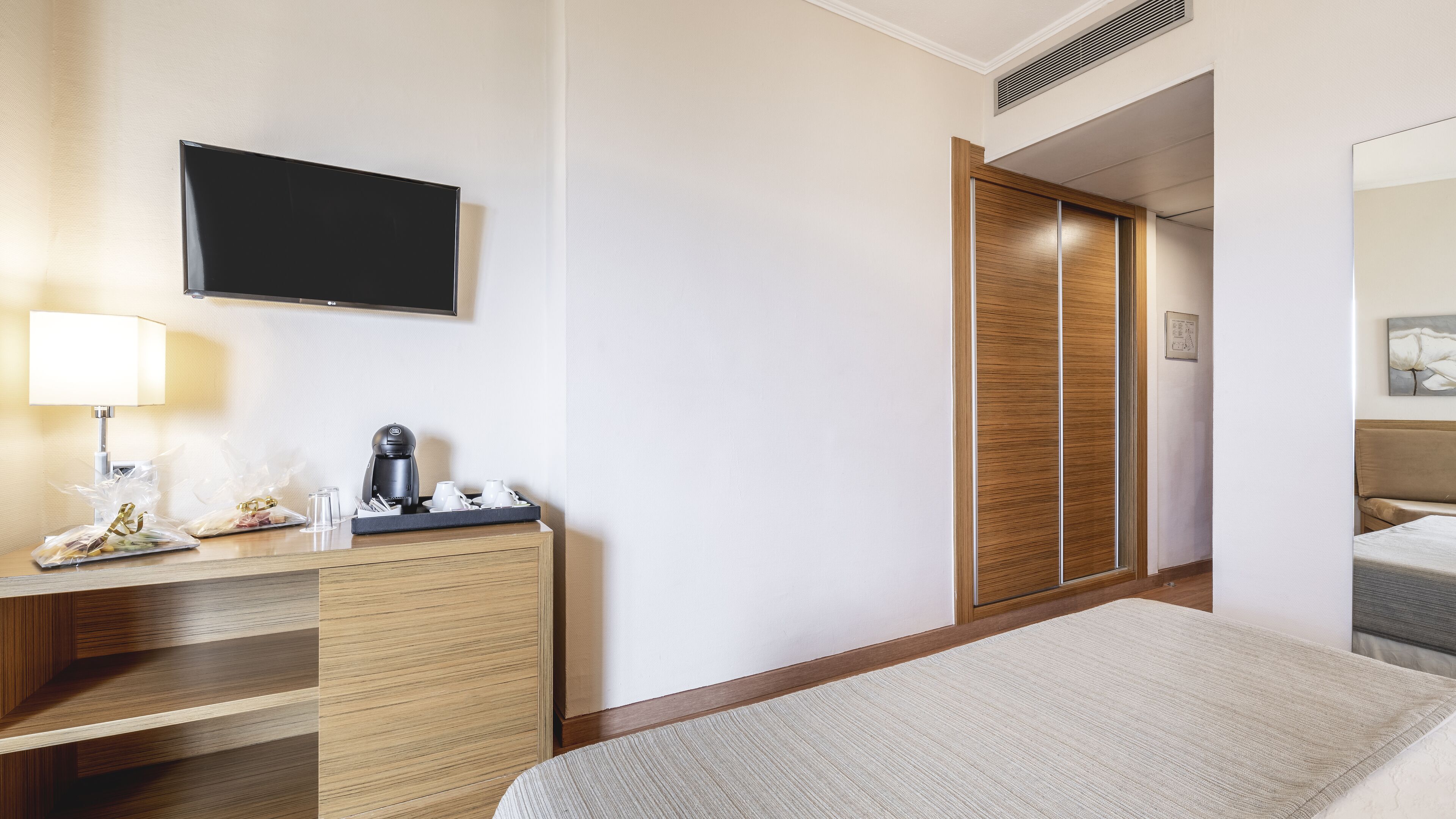 Habitación de hotel con cama, escritorio con lámpara, cafetera y televisor en la pared.