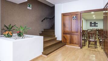 Un área de recepción con mostrador blanco, plantas en macetas, una escalera y una puerta de madera con un cartel.