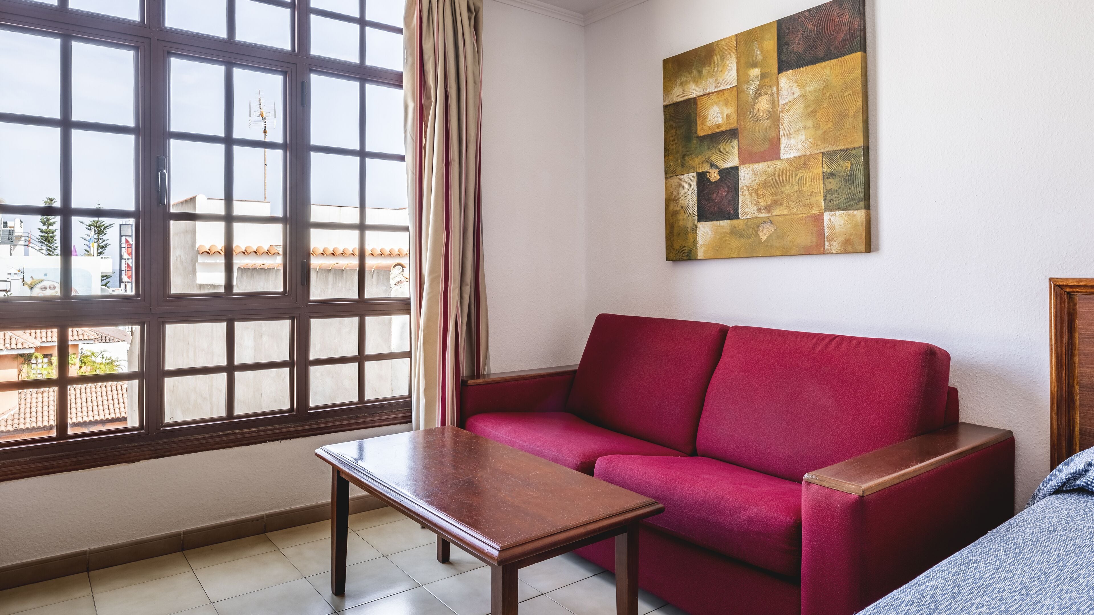A living room with a red sofa, a wooden coffee table, a large window with curtains, and a wall-mounted abstract painting.