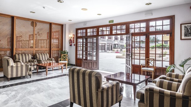 Un vestíbulo con sillones a rayas, una mesa de centro de madera y vistas a la calle a través de grandes puertas de madera.