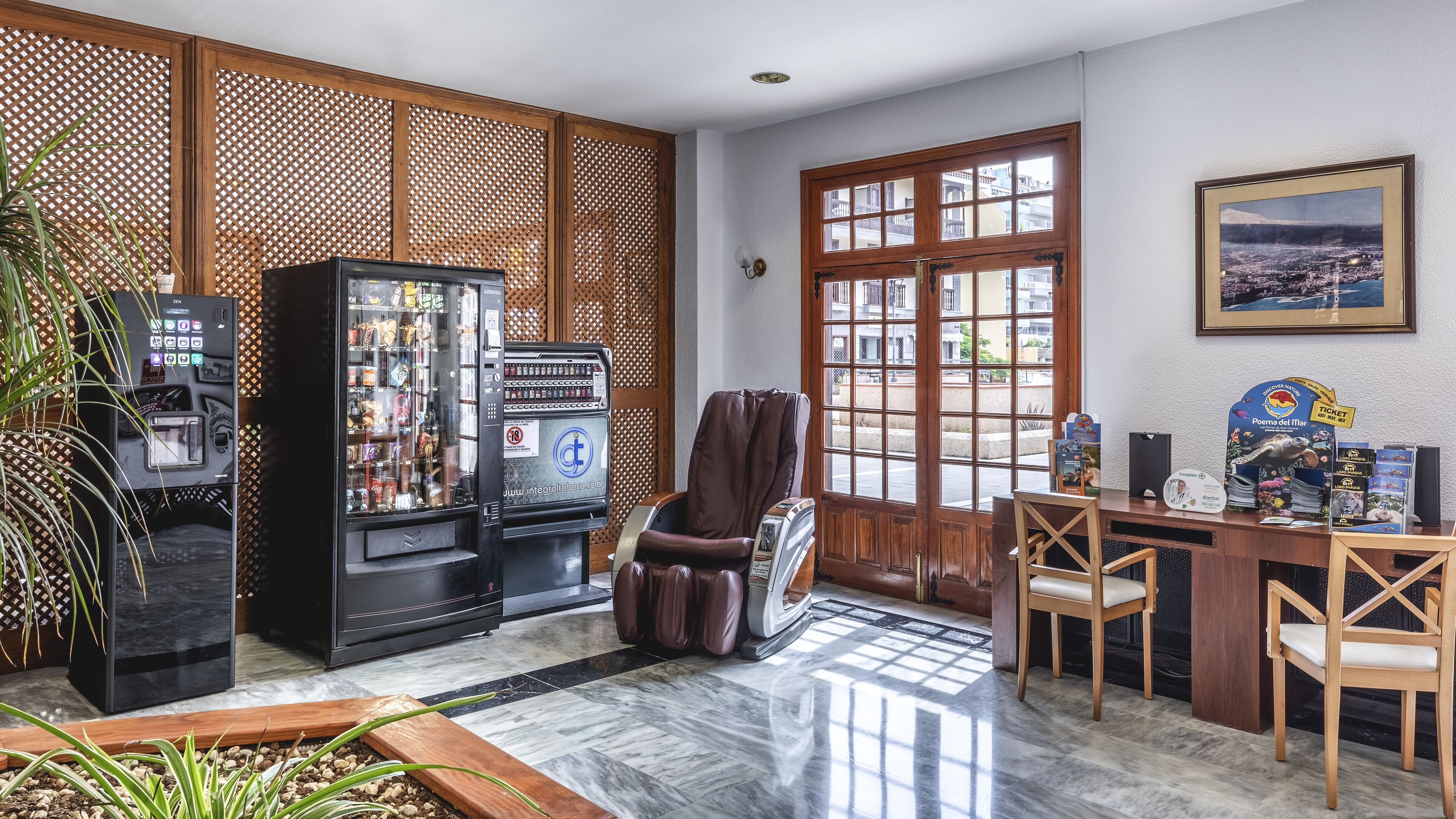 A room with a massage chair, vending machines, a wooden desk, chairs, and a large window.