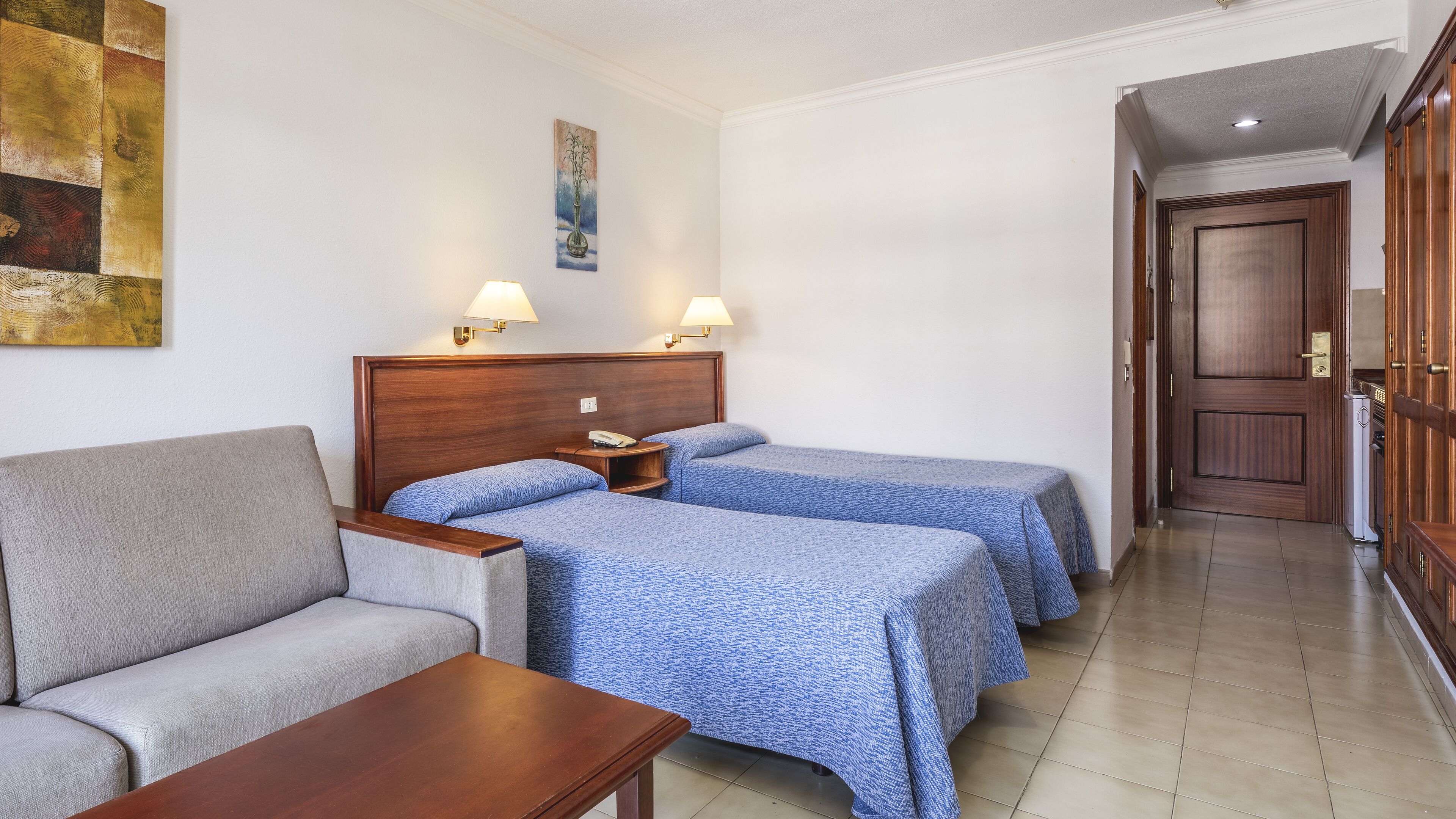 A hotel room with a sofa, two single beds with blue bedding, a nightstand with a lamp, a painting on the wall, and a wooden door.