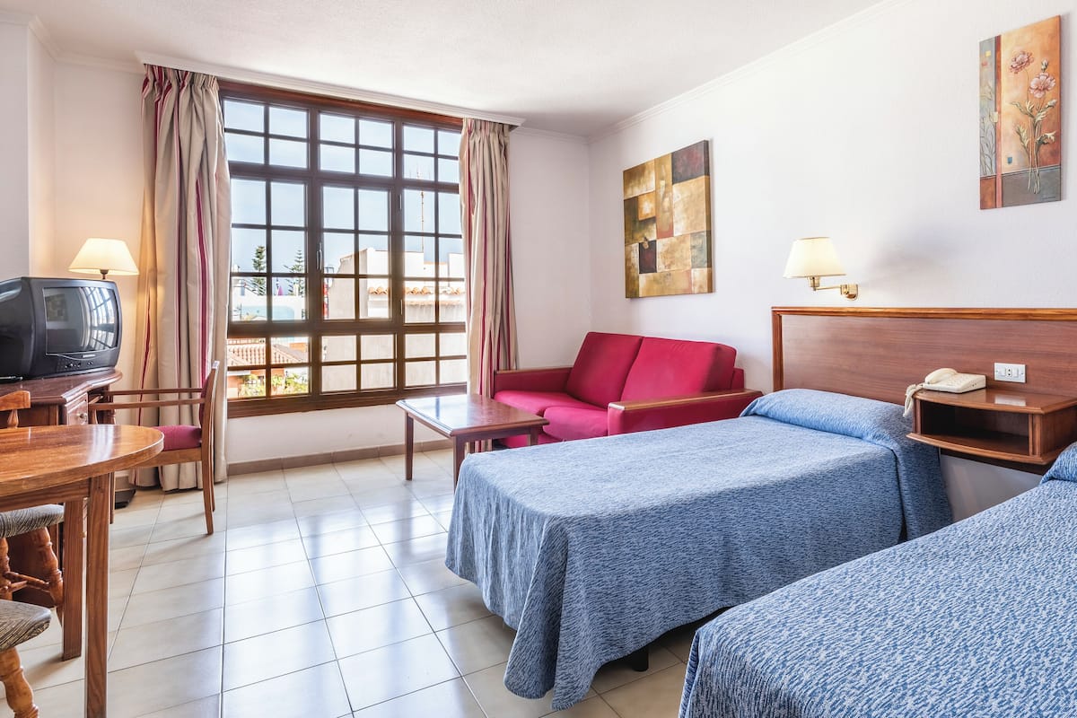 A hotel room with two beds, a red sofa, a wooden desk with a television, and a window with curtains.