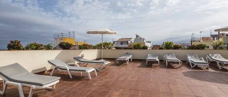 A rooftop terrace with multiple sun loungers and a single umbrella, surrounded by terracotta tiles and a view of the cityscape and mountains.