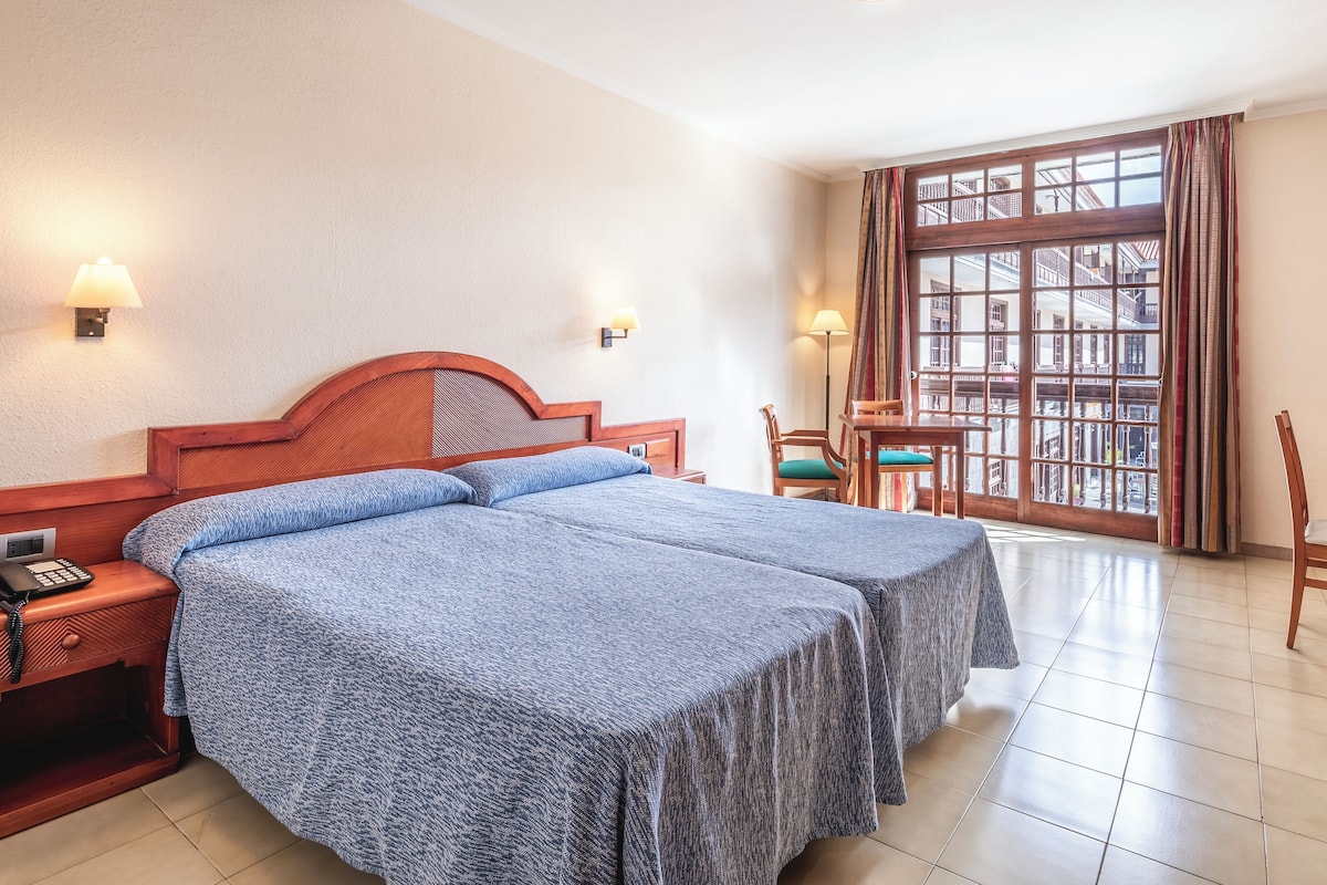 A hotel room with a double bed, bedside lamps, a desk with a television, and a window with a view of the city.
