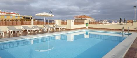 A swimming pool with lounge chairs and umbrellas, surrounded by a fenced area with buildings in the background.