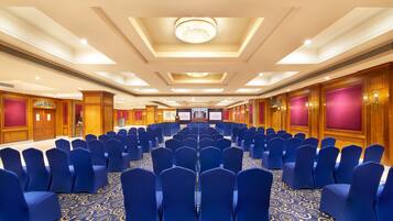 Un salón de conferencias con filas de sillas azules, una alfombra estampada y paneles de madera en las paredes.