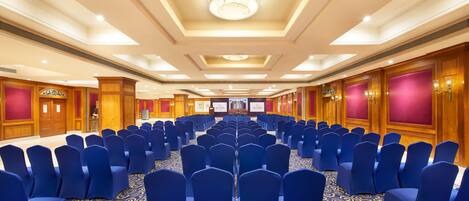 A conference room with rows of blue chairs, a patterned carpet, and wooden paneling on the walls.